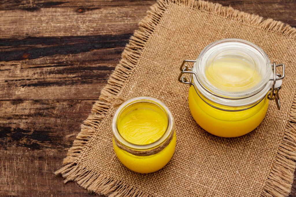 pure or desi ghee (ghi), clarified melted butter. healthy fats bulletproof diet concept or paleo style plan. glass jar, silver spoon on vintage sackcloth. wooden boards background